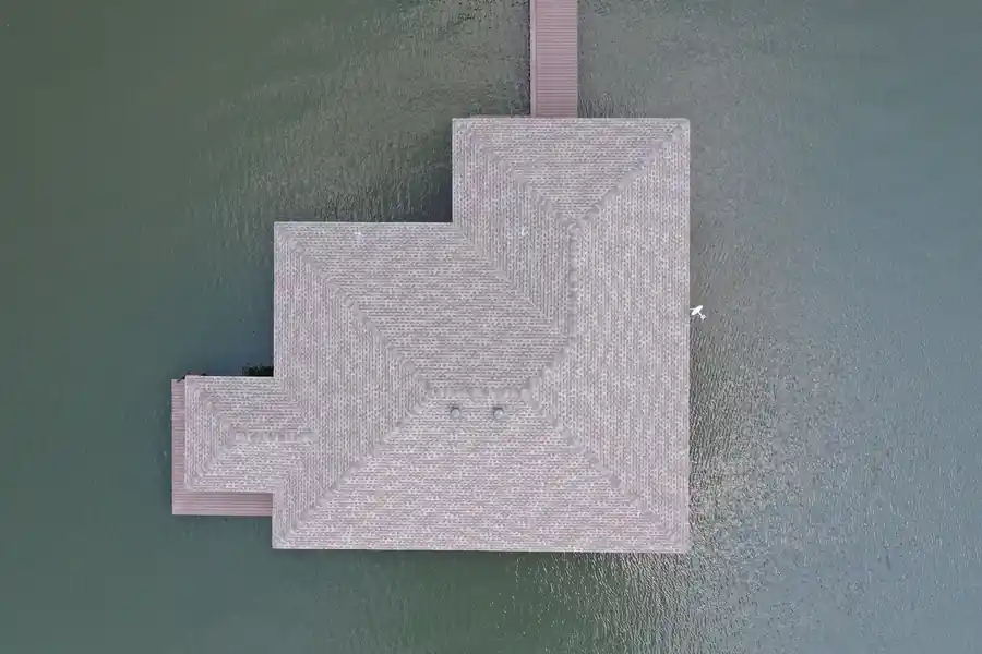 Straight-down drone photo of a shingled boathouse roof standing over open lake water, a pier running to the shore