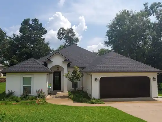 Street view of a white stucco home with a dark hip roof under a cloudy blue sky